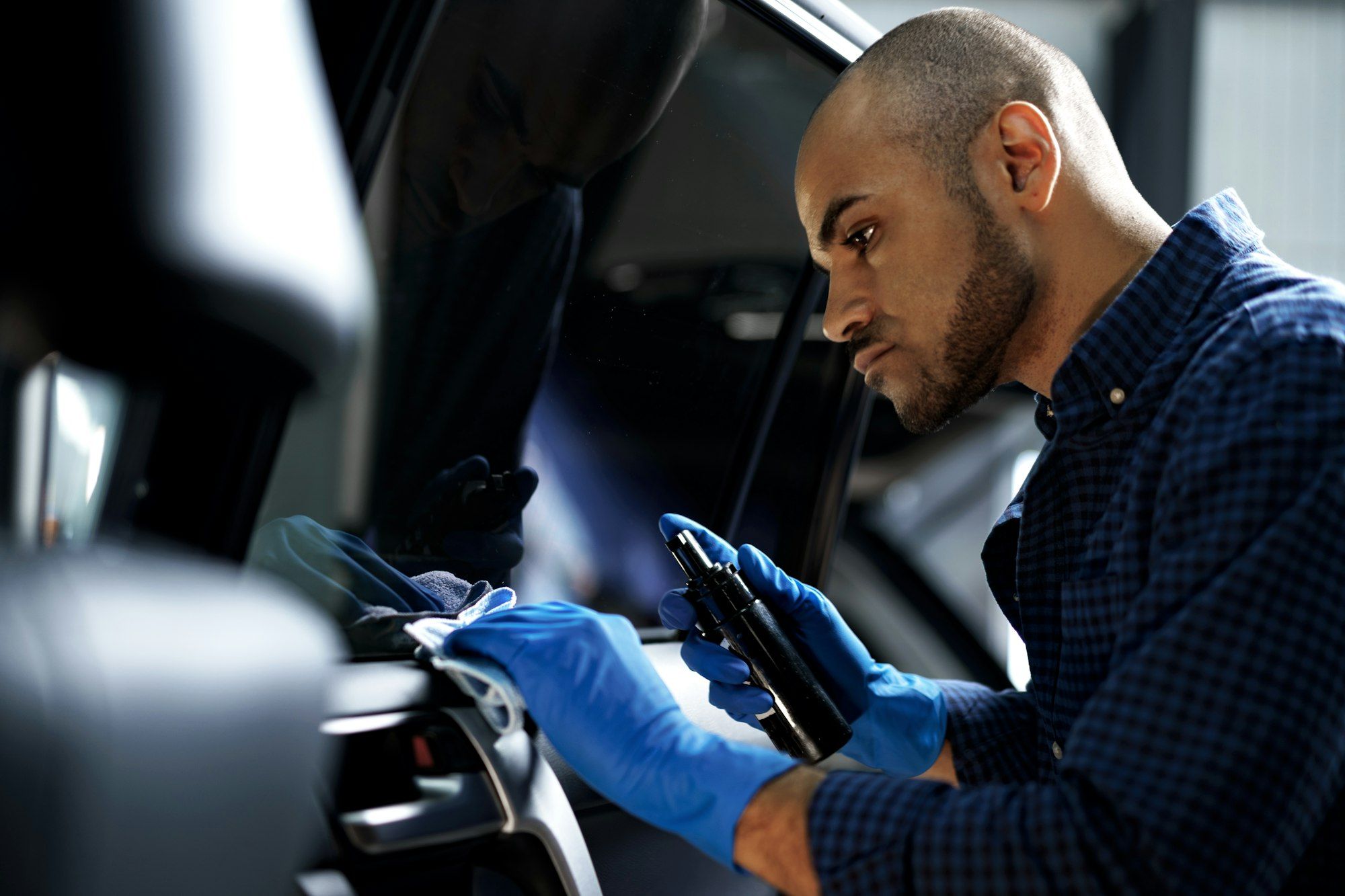 Eine Person mit blauen Handschuhen und einem blau karierten Hemd reinigt sorgfältig den Innenraum eines Autos und bereitet es möglicherweise für eine Kfz-Gutachten-Prüfung in Berlin vor. Sie hält eine Sprühflasche in der Hand und wischt eine Oberfläche mit einem Tuch ab, wobei sie sich intensiv auf ihre Aufgabe konzentriert.