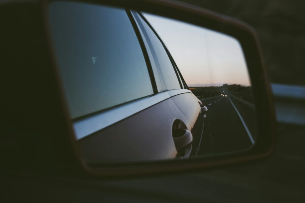 Der Seitenspiegel eines Autos spiegelt den Blick auf eine Straße in der Abenddämmerung wider, in der Ferne nähert sich ein anderes Fahrzeug. Der Himmel ist schwach beleuchtet und deutet auf das Ende des Tages hin. Das elegante Äußere des Autos ist teilweise im Rahmen des Spiegels sichtbar.