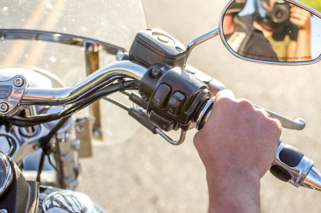 Nahaufnahme der Hand einer Person, die den Lenker eines Motorrads hält. Die Bedienelemente am Lenker sind deutlich zu erkennen, und im Rückspiegel spiegelt sich ein Fotograf, der das Bild macht. Im Hintergrund ist eine verschwommene Straße zu sehen.