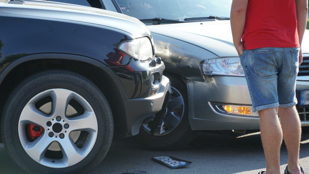 Ein Mann in rotem Hemd und Jeansshorts steht neben zwei Autos, die in einen leichten Unfall verwickelt waren. Die Frontpartien beider Autos sind beschädigt, dazwischen liegt ein kaputtes Nummernschild auf dem Boden. Die Szene spielt an einem sonnigen Tag.