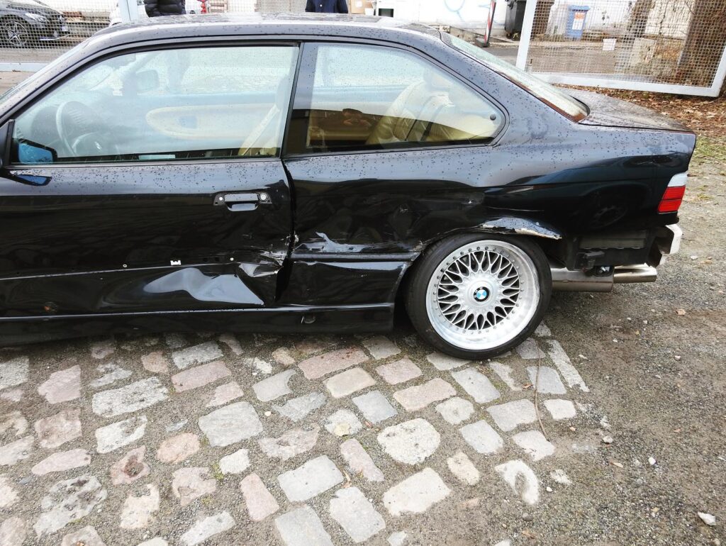 Eine schwarze Limousine mit erheblichen Schäden an der hinteren Beifahrertür und am Kotflügel, geparkt auf einem gepflasterten Platz. Das Hinterrad ist freiliegend und sichtbar verbogen, und der Lack des Autos ist zerkratzt und voller Dellen. Der Innenraum des Autos ist durch die Fenster zu sehen, was deutlich macht, dass ein Unfallgutachten erforderlich ist.